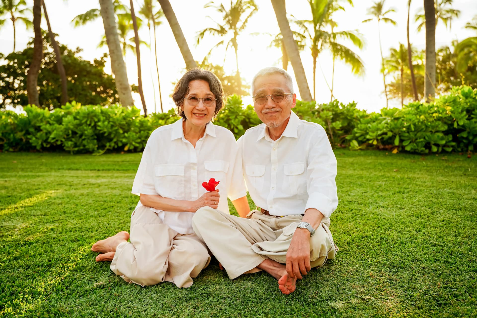 Elderly couple sitting on grass surrounded by palm trees at sunset